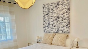 A neatly made bed with a patterned bedspread, two bedside tables with lamps, a wall-mounted artwork, and a window with sheer curtains. - Apartamento 2 (Peníscola)