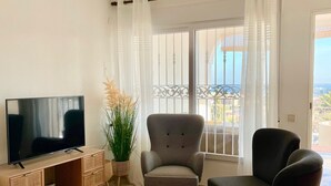 A living room with a TV, two armchairs, a wooden TV stand, and a large window with curtains. - Apartamento 2 (Peníscola)
