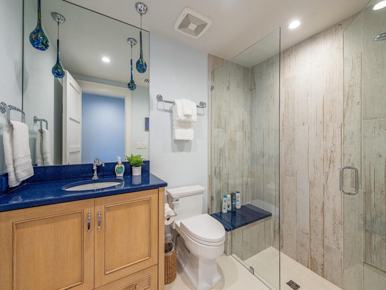 Un baño moderno con encimera azul, armarios de madera, inodoro blanco y cabina de ducha con mampara de cristal.