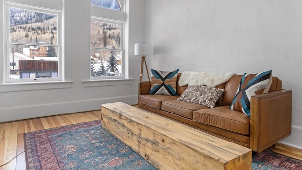 A room with a brown leather sofa, a wooden bench, and a patterned rug. There are two windows with a view of a snowy mountain and buildings. - Lawless: A Boutique Basecamp in Rico (Rico)