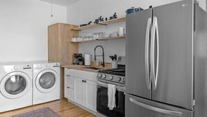 A compact kitchen with a stainless steel refrigerator, a built-in oven, a washing machine, and wooden shelves above the counter. - Lawless Rico: Enterprise Suite (Rico)