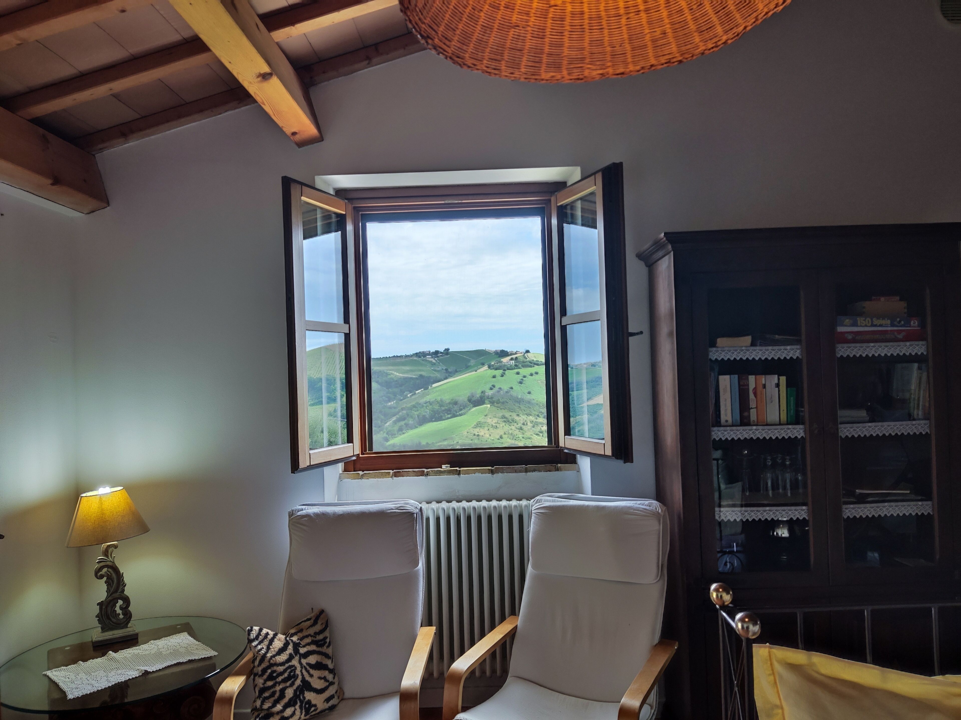A room with a large window, a radiator, a chair, a table with a lamp, and a bookshelf.