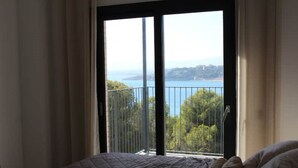 Ein Schlafzimmer mit großem Fenster und Balkonblick auf Bäume und ein Gewässer.