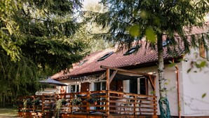 Une cabane en bois avec un toit recouvert de tuiles rouges, une véranda dotée de balustrades en bois et un jardin agrémenté de plantes et d’un banc.