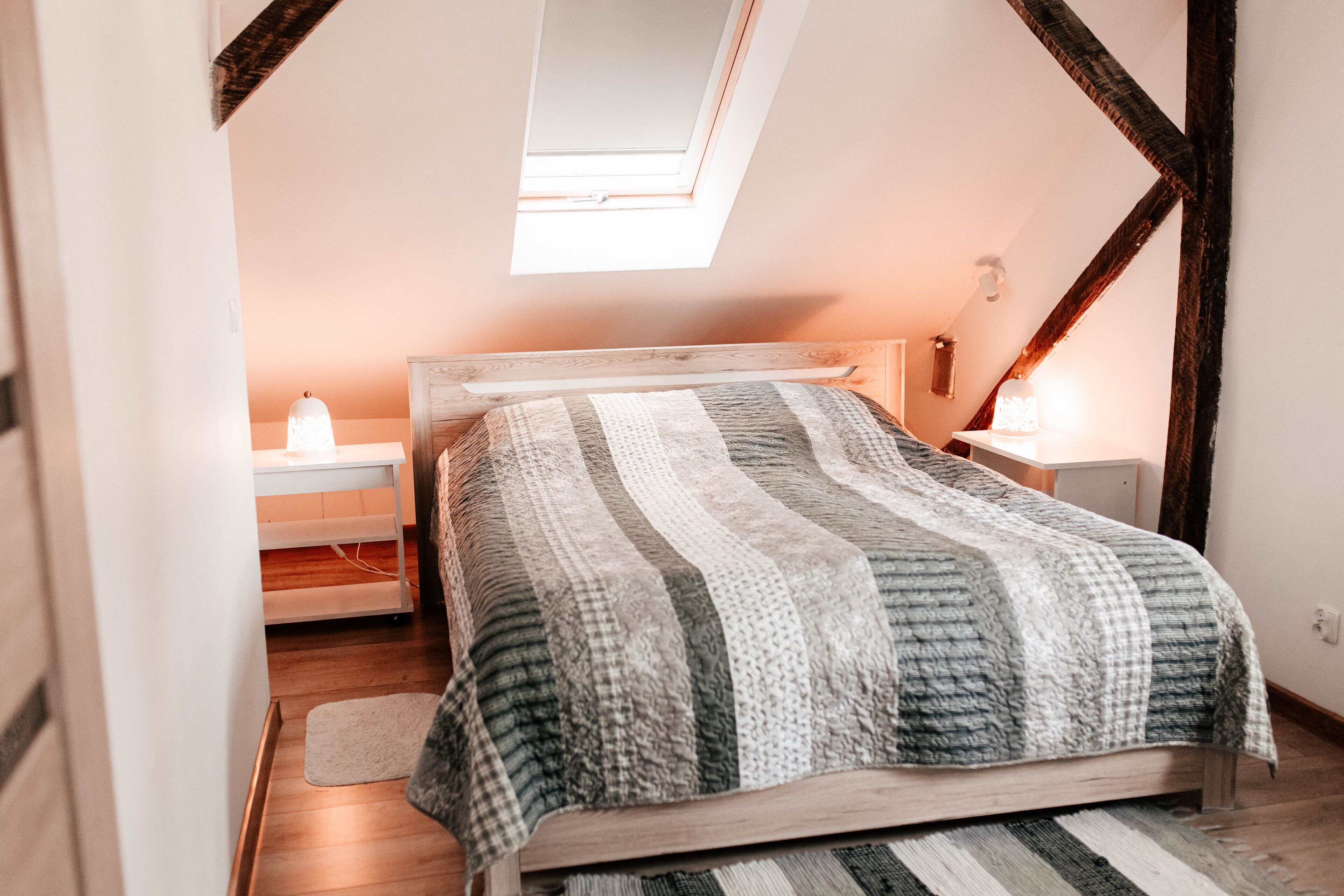 A bedroom with a sloped ceiling, a skylight, a wooden bed frame, a striped blanket, bedside tables with lamps, and a small rug.