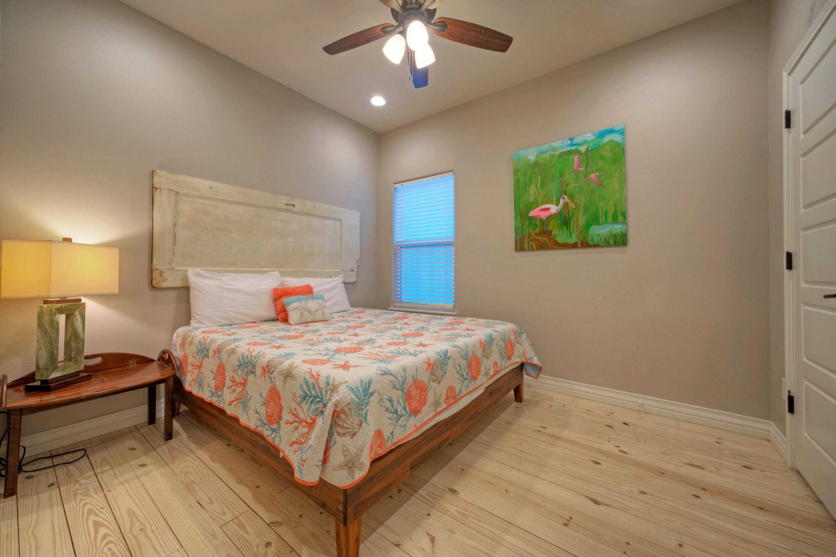 Un dormitorio con cama de madera, una mesita de noche, ventilador de techo, ventana con persianas y un cuadro en la pared.