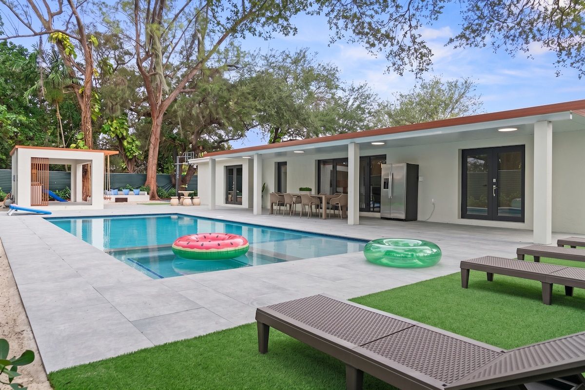 A modern outdoor pool area with a covered seating area, lounge chairs, and a small structure.