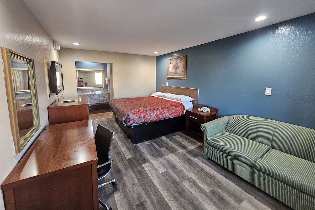 A hotel room with a bed, a desk, a chair, a green sofa, a mirror, and a television.