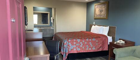 A hotel room with a bed, a desk, a chair, a nightstand, a phone, and a painting on the wall.