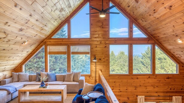 Ein gemütliches Blockhaushaus-Interieur mit schrägen Wänden, großen Fenstern, einem Deckenventilator und Holzboden.