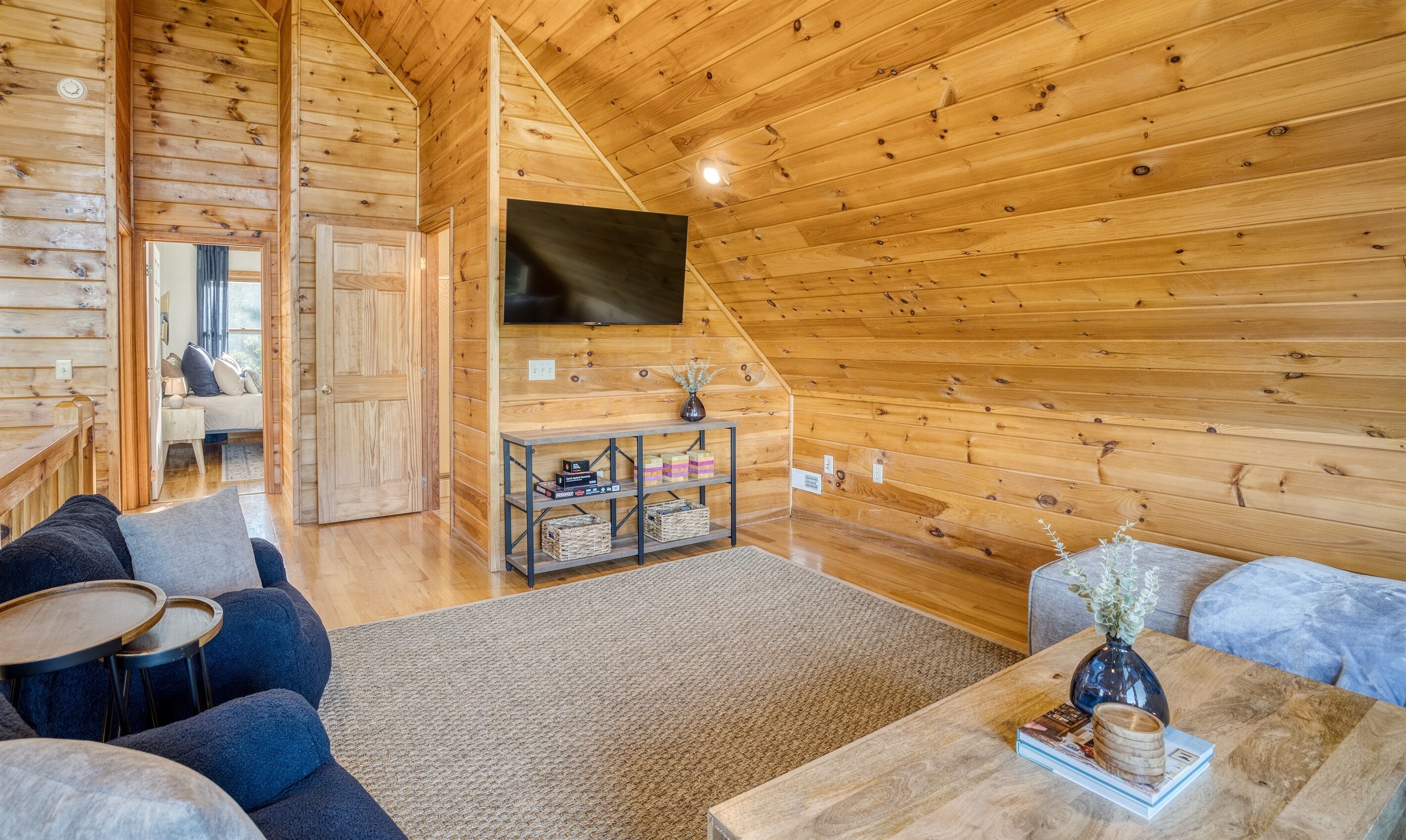 Een gezellig houten blokhutinterieur met een aan de muur bevestigde televisie, een comfortabele zithoek met een bank en fauteuils, en een kleine tafel met decoratieve objecten.