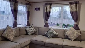 A corner living room with a beige sectional sofa, patterned cushions, and a window with purple curtains. - Purple Palace, 3-bed Caravan, Ingoldmells (Skegness)