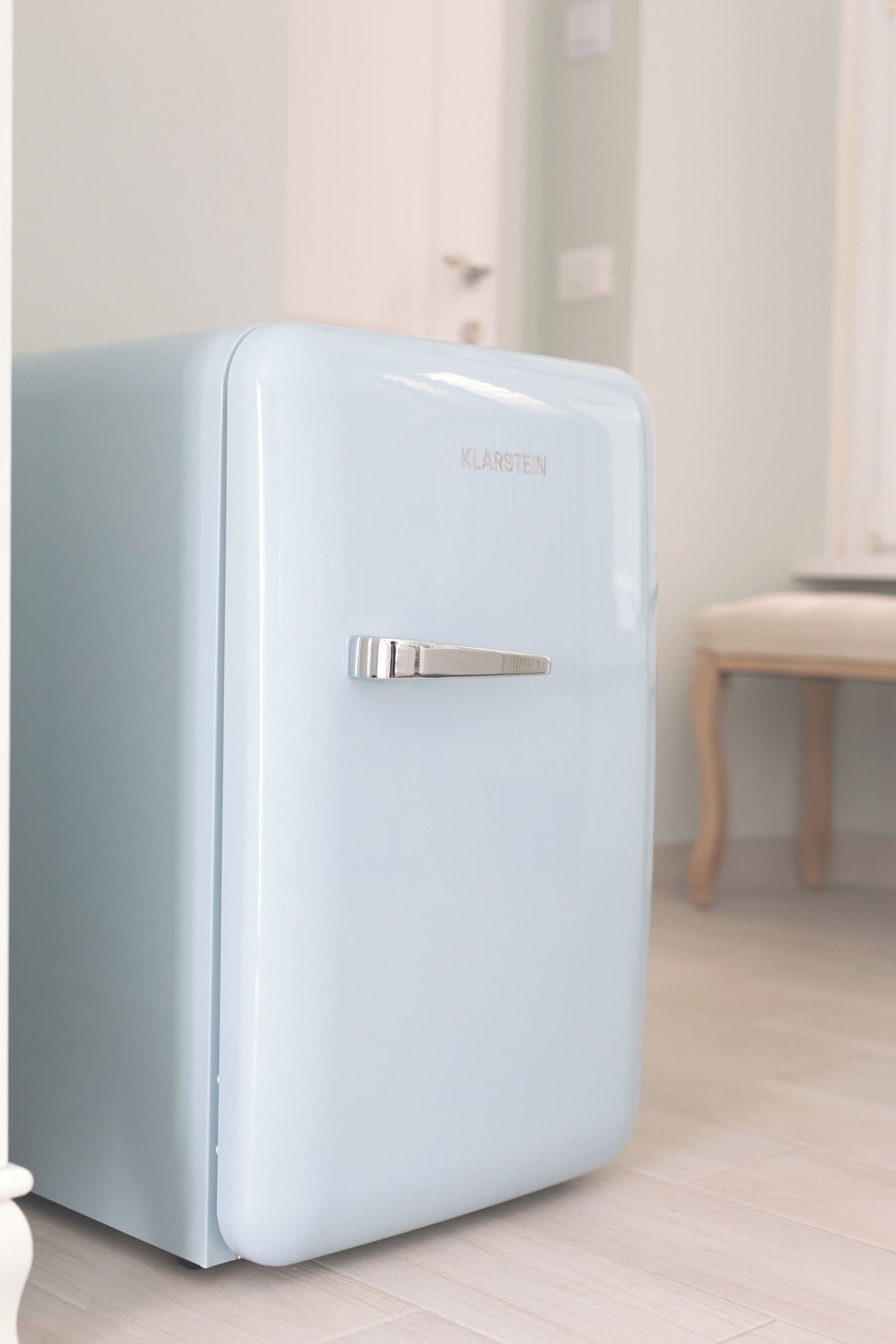 Un réfrigérateur de style rétro, de couleur bleu clair, doté d’une poignée, est placé dans une pièce au plancher en bois, avec une chaise visible au fond.