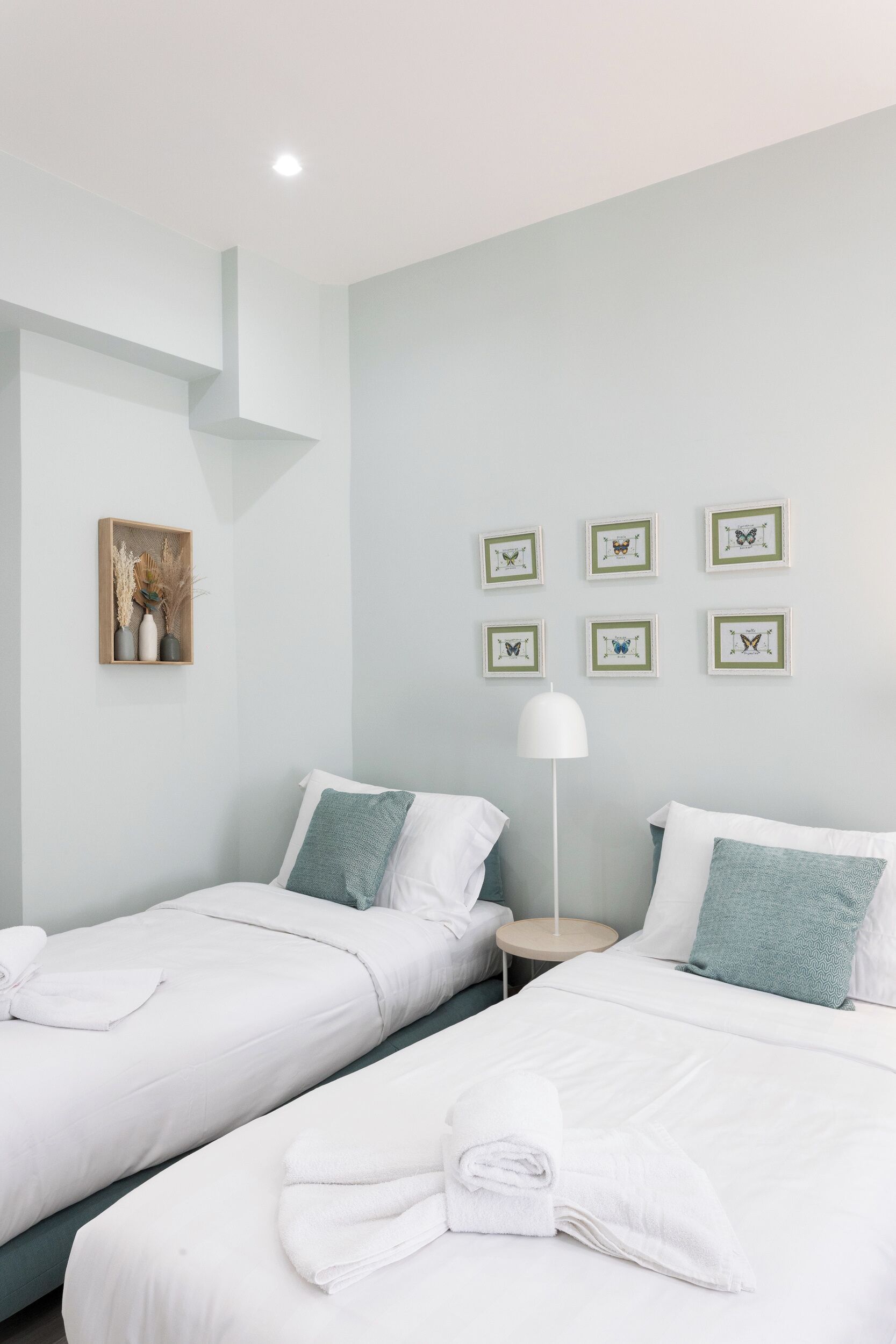 A hotel room with two beds, white bedding, a small bedside table with a lamp, and framed butterfly artwork on the wall.