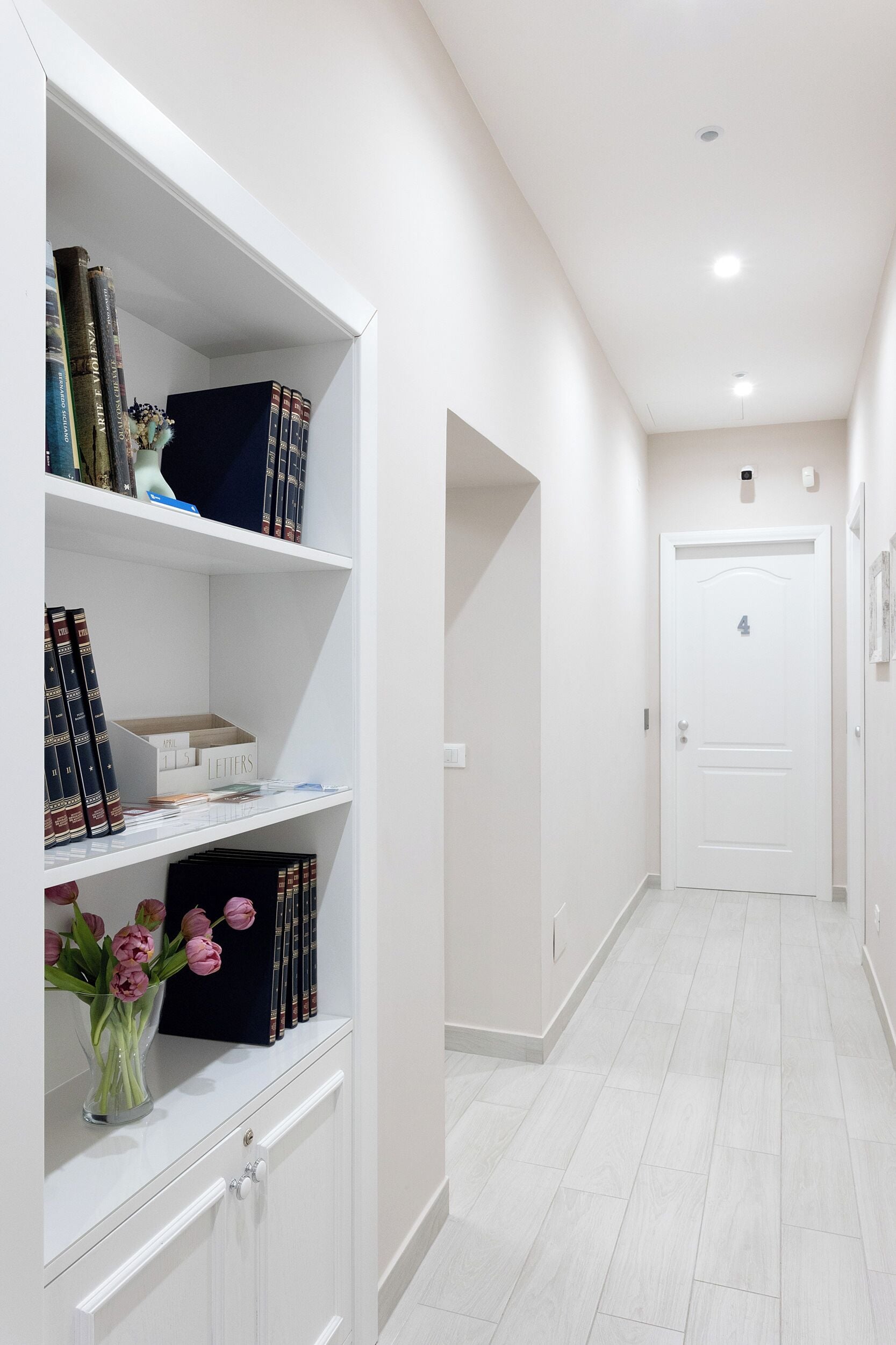 Un couloir avec une bibliothèque blanche, une porte portant l’inscription « 4 », et un vase rempli de fleurs roses.
