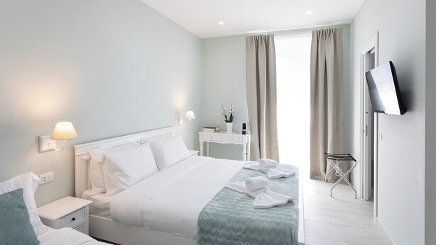 A hotel room with a neatly made bed, a bedside table with a lamp, a small desk with a chair, a TV mounted on the wall, and a window with curtains. - Colosseo House (Rome)