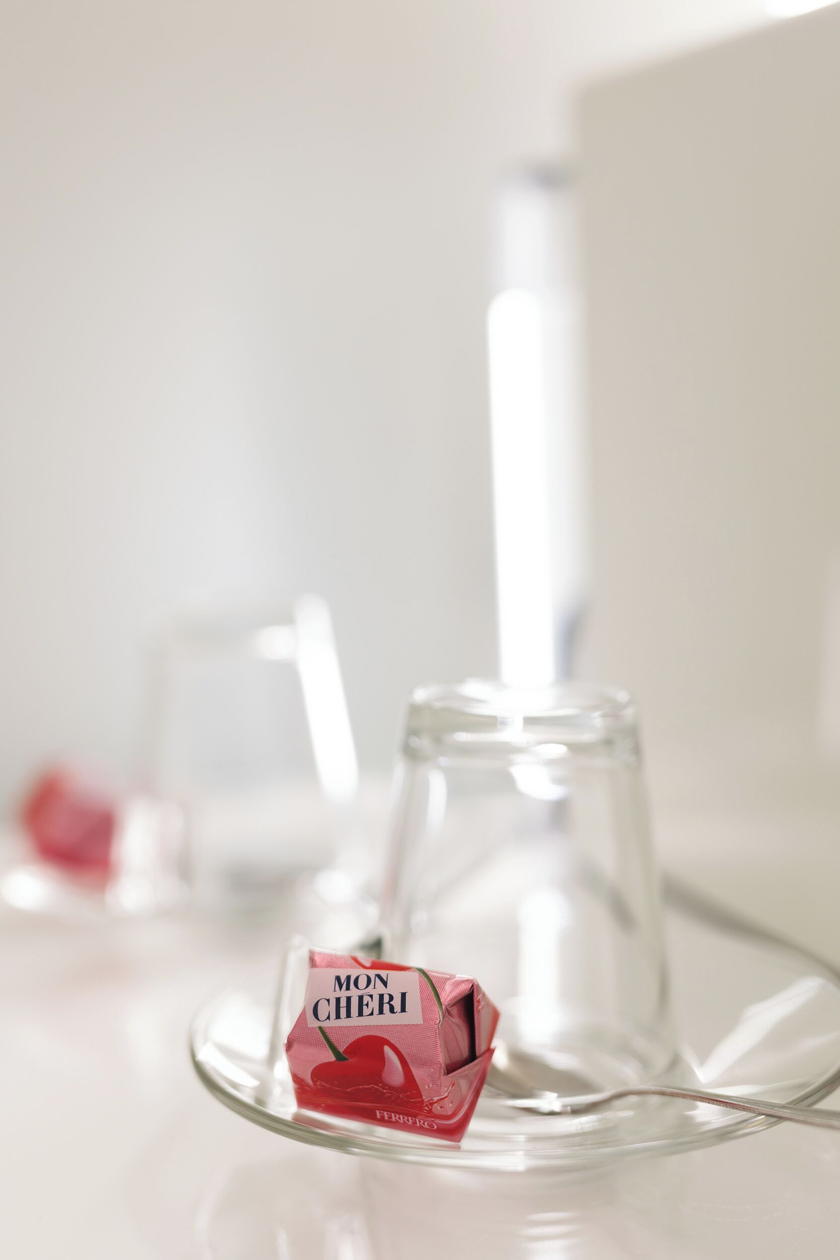 A clear glass wine glass on a reflective surface with a small packet of Mon Cheri chocolate.