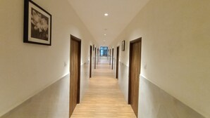 Un long couloir étroit, avec des portes en bois de chaque côté, un tableau encadré sur le mur de gauche et un plancher de couleur claire.