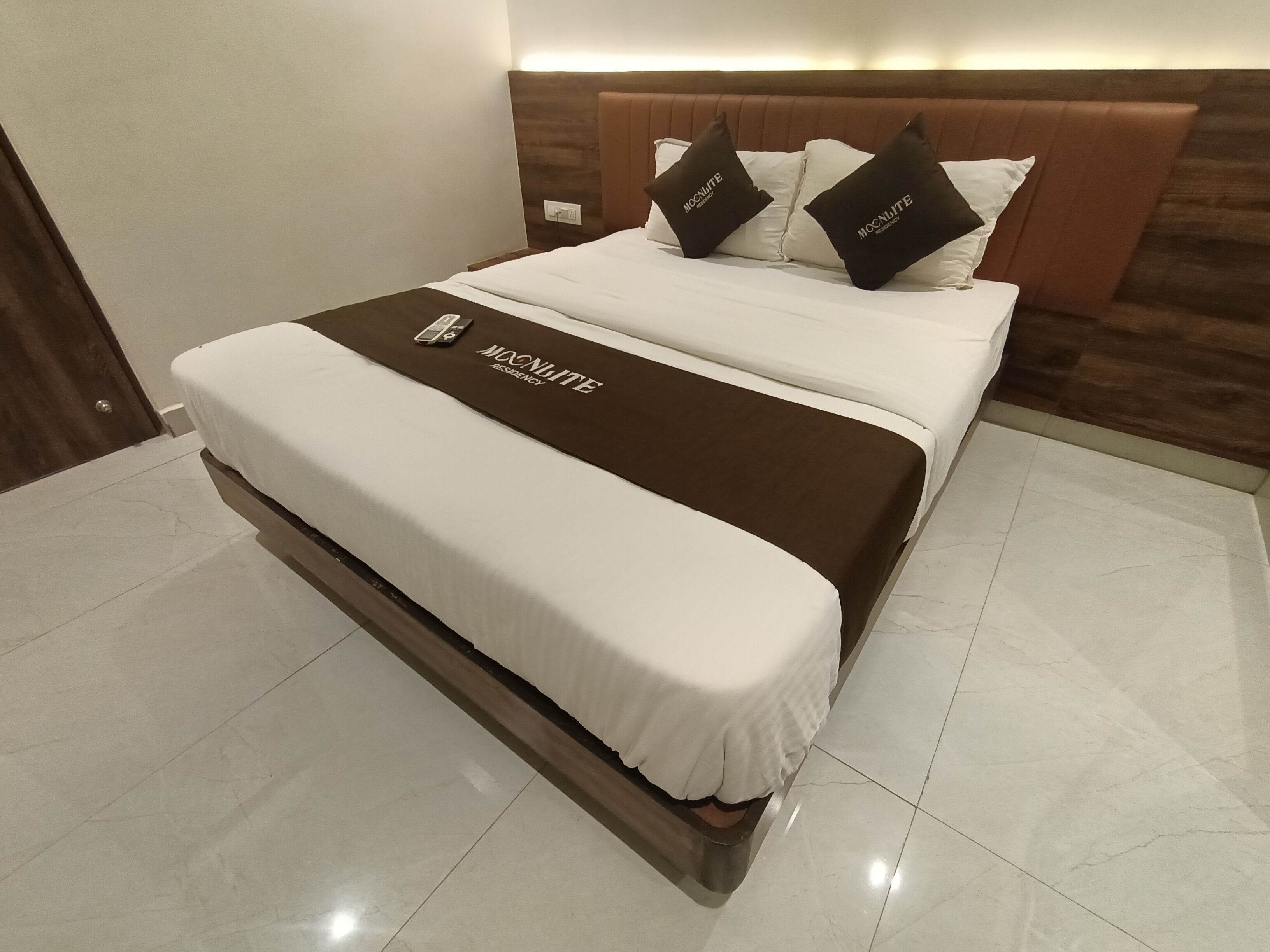 A hotel room with a bed, a brown headboard, and a white bedspread with a logo.
