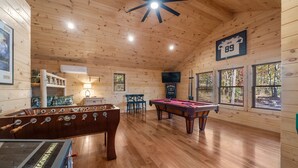 Une pièce au plancher de bois, équipée d’un baby-foot, d’une table de billard et d’un ventilateur de plafond.