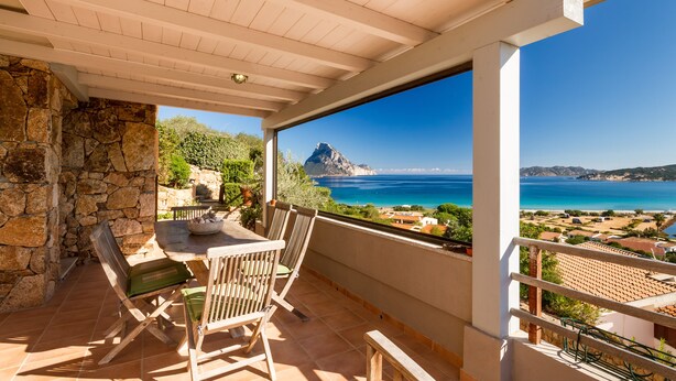 A balcony with a stone wall, wooden furniture, and a view of the sea and mountains.