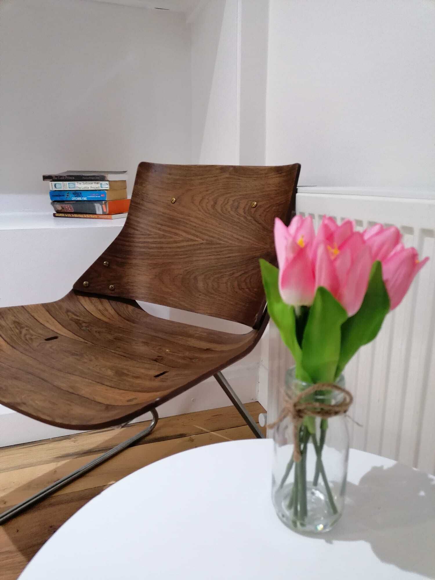 Ein Holzhocker mit Metallnieten, ein weißer Tisch und eine Vase mit rosa Tulpen.