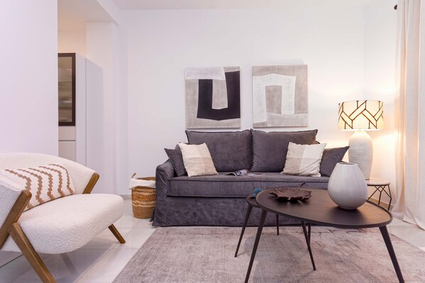 A modern living room with a grey sofa, a white armchair, and a small table with a vase. There are abstract paintings on the wall and a lamp on the table.
