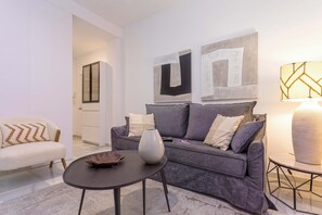 Una sala de estar moderna con un sofá gris, una mesita redonda y un jarrón decorativo. Hay pinturas abstractas en la pared y una lámpara sobre una mesita auxiliar.