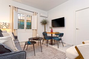 Una sala moderna con sofá, una mesa de centro, una mesa de comedor con sillas, un televisor montado en la pared y una ventana con cortinas.