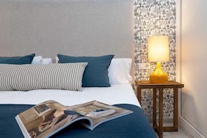 A neatly made bed with blue and white bedding, a wooden nightstand with a yellow lamp, and a magazine on the bed.