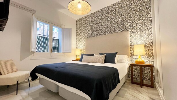 Een slaapkamer met een groot bed, een raam, een behang met patroon en een klein tafeltje met een lamp.