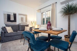 A compact living space with a sofa, dining table, and chairs. There is a window with curtains, a potted plant, and abstract wall art. - Genteel Home Baños IV (Seville)