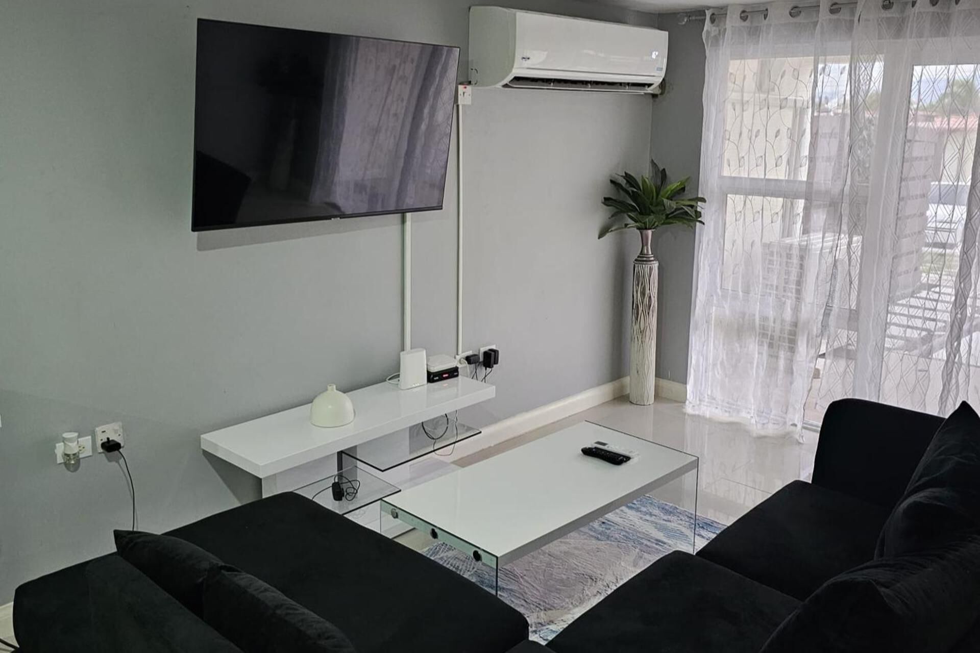 A modern living room with a flat-screen TV, air conditioning unit, white console table, and black sofas.