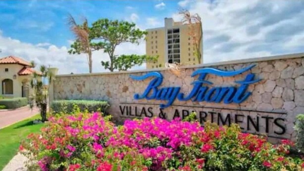 A sign for 'Bayfront Villas & Apartments' with a stone wall and colorful flowers in the foreground. - Apartment Sleeps 6 With Pool Access (Portmore)
