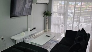 A modern living room with a flat-screen TV, air conditioning unit, and a large window with sheer curtains. - Apartment Sleeps 6 With Pool Access (Portmore)