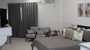 Quarto de hotel com cama, escrivaninha, cadeira, mesinha redonda e televisão.