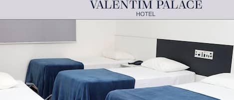 Habitación de hotel con tres camas, cada una cubierta con una colcha azul. La habitación tiene un diseño moderno, una pared blanca y un cabecero negro.