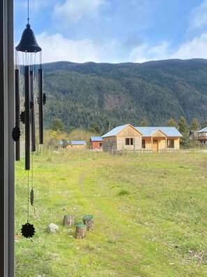 On aperçoit une cabane rustique avec un toit en métal, entourée d’un champ herbeux et, au loin, d’une chaîne de montagnes boisée.