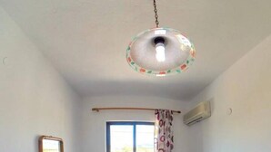 A small, single-bed room with a wooden bed frame, a blue bedspread, a hanging light fixture, a window with curtains, and an air conditioning unit on the wall. - Blue House Beach Apartment - Sleeps 4 & Balcony (Rhodes)