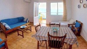 A cozy living room with a blue sofa, a wooden dining table with a checkered tablecloth, and a view of the sea through the open door. - Blue House Beach Apartment - Sleeps 4 & Balcony (Rhodes)