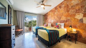A room with two beds, a ceiling fan, a stone wall, a nightstand, and a view of the outdoors. - Sueño - Playacar 2 Rosa Blanca - 2 BR (Playa del Carmen)