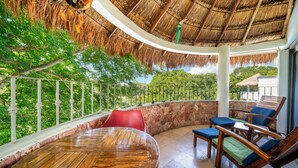 A wooden table and chairs in a covered outdoor area with a thatched roof, surrounded by stone walls and a view of lush greenery. - Sueño - Playacar 2 Rosa Blanca - 2 BR (Playa del Carmen)