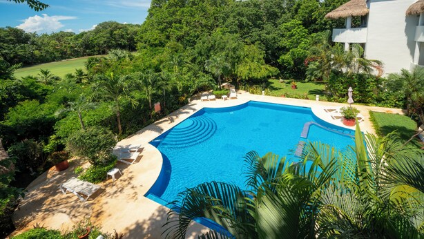A pool surrounded by lush greenery and a white building with a thatched roof. - Sueño - Playacar 2 Rosa Blanca - 2 BR (Playa del Carmen)