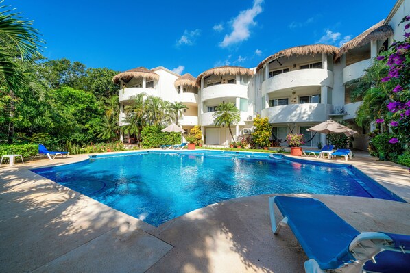 A swimming pool with lounge chairs, surrounded by multi-story buildings with thatched roofs, and lush greenery.