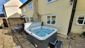 A hot tub in a backyard with stone flooring, a table and chairs, and a stone wall.