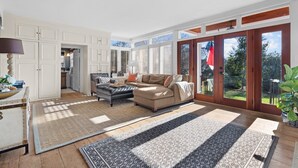 A spacious living room with a large sectional sofa, a coffee table, and a dining area with a table and chairs. There are large windows with blinds, a potted plant, and a view of the outdoors.