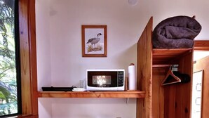 Una cocina compacta con microondas, cafetera y tostadora. Hay un cubo de basura pequeño y un armario de madera con un objeto colgando.