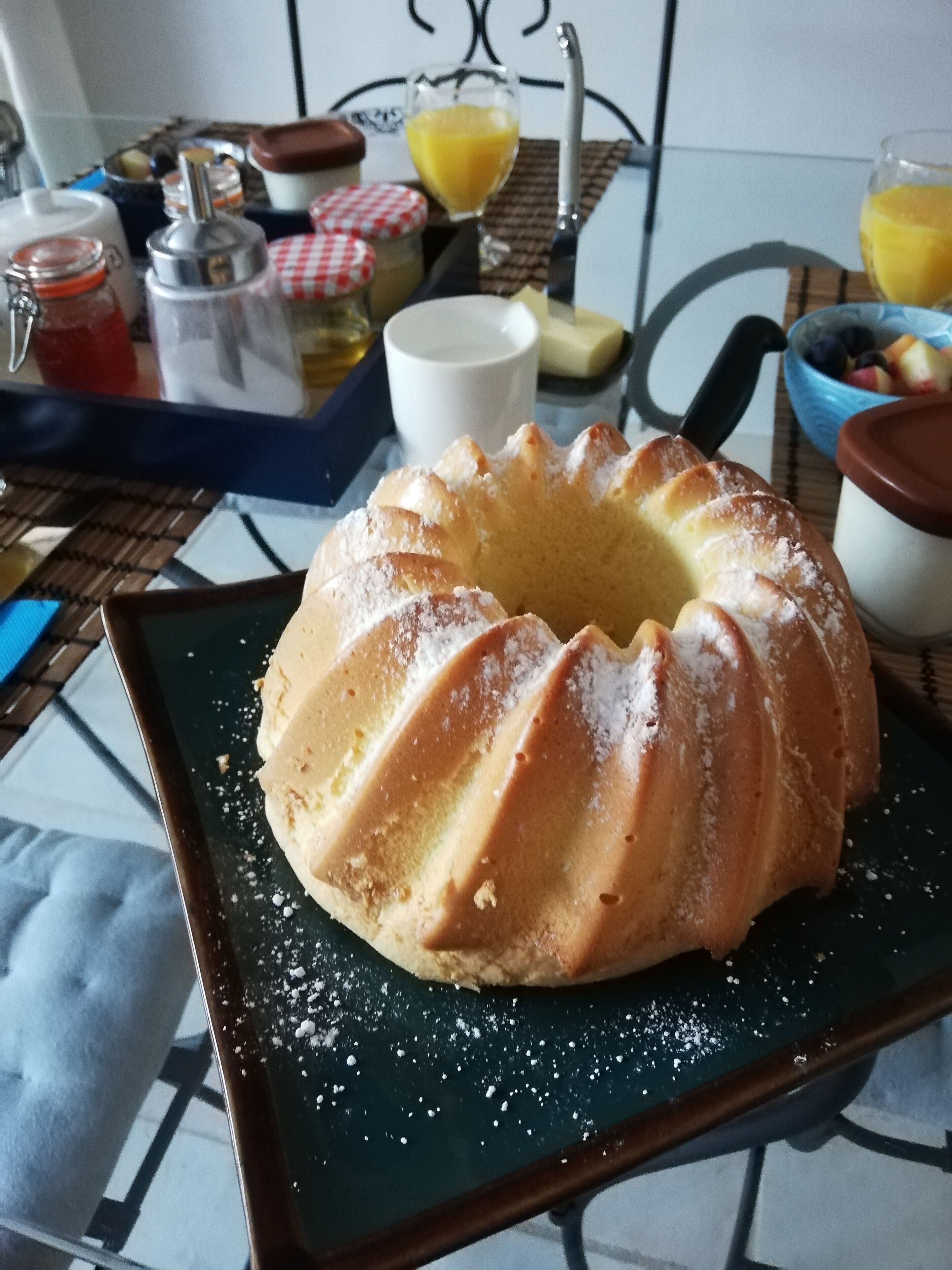 Un pastel Bundt en un plato con azúcar glas, una taza de café, un vaso de zumo de naranja y un bol de fruta sobre una mesa.