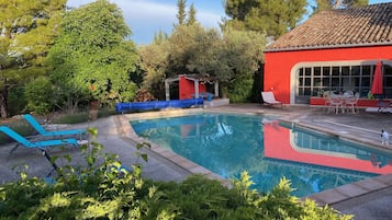 En swimmingpool med blå liggestole, en rød bygning med store vinduer og grøn bevoksning omkring.