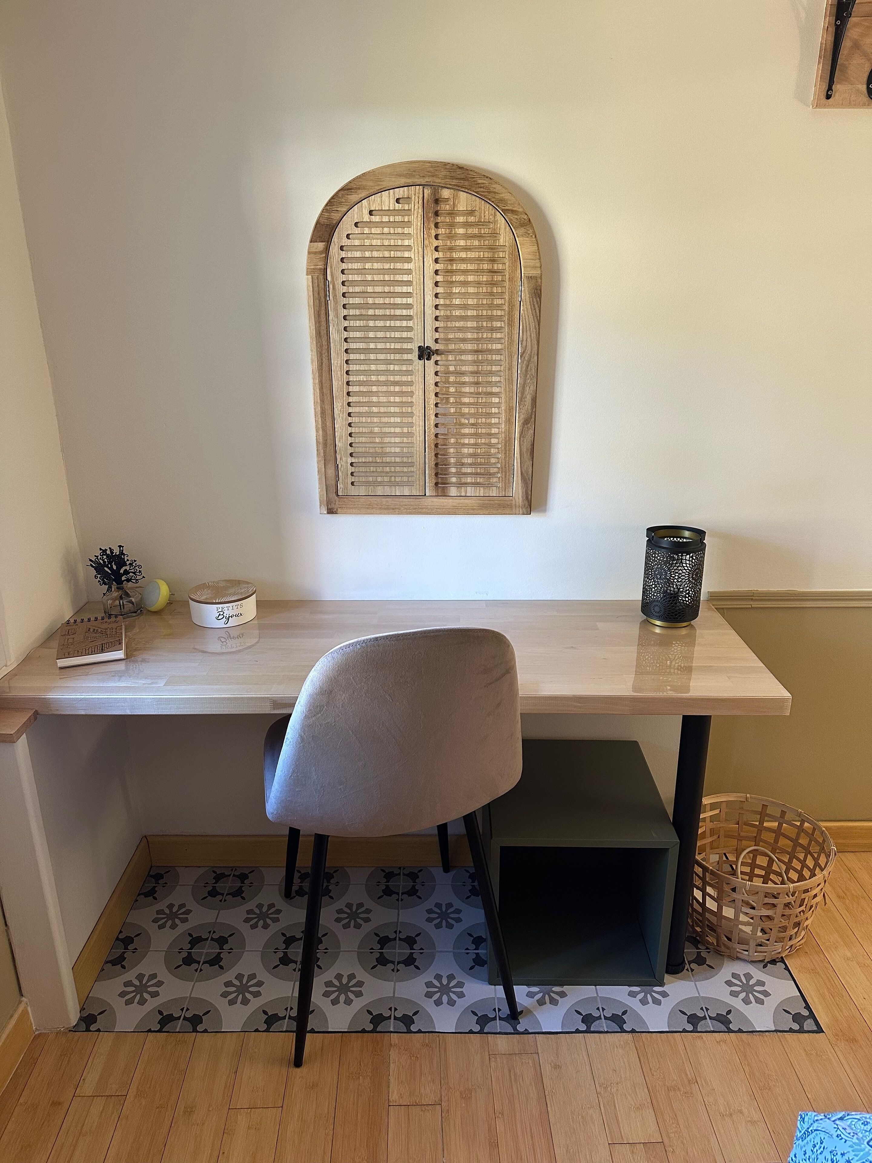 A study with a wooden desk, a chair, a window with shutters, a small table with decorative items, and a woven basket.